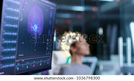 Woman Wearing Brainwave Scanning Headset Sits in a Chair In the Modern Brain Study Laboratory/ Neurological Research Center. Monitors Show EEG Reading and Brain Model.