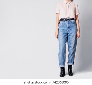 Woman Wearing Casual Outfit With White Shirt, Blue Vintage High-waisted Mom Jeans With Black Ankle Boots And Black Belt Isolated On Grey Background. Copy Space