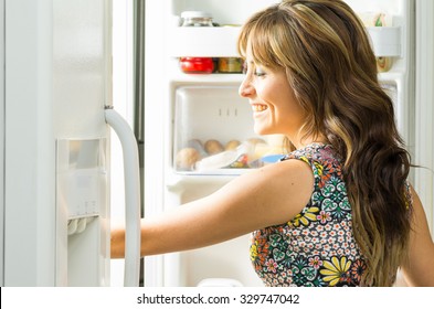 Woman wearing colorful dress in modern kitchen opening fridge door and reaching inside.
