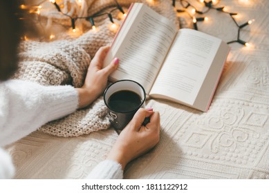 Woman Wearing Cozy Sweater,  Holding Mug Of Warm Coffee And Reading Book In Bed On Cold Autumn Or Winter Day. Relaxation And Hygge Concept