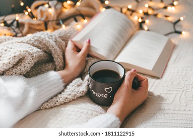 Woman Wearing Cozy Sweater,  Holding Mug Of Warm Coffee And Reading Book In Bed On Cold Autumn Or Winter Day. Relaxation And Hygge Concept