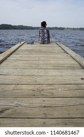 A Woman Wearing Dark Clothes And Dark Curly Hair In A Bun Is Sitting Alone Next To A Pair Of Shoes On A Long Wooden Pier And Looking Ahead Into The River On A Sunny Day With Cloudy Blue Sky.