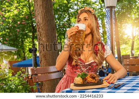 Foto Bild Frau im Dirndl im bayerischen Biergarten sitzend und Bier trinkend mit traditioneller bayerischer Küche mit Schweinshaxe, gebratener Schweinshaxe auf einem Tisch