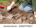 A woman wearing dirty pants and rubber gloves holds an excavating trowel digging for artifacts. The archaeologist is sweeping soil from rocks uncovering pottery and bones as part of research.