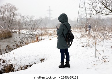 Woman wearing down jacket standing with smartphone in hands river coast at snowy weather. Leisure in winter park, cold season