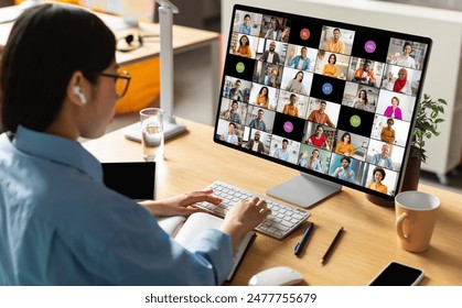 A woman wearing earbuds sits at her desk in front of a computer monitor displaying a video conference call with many participants. She is actively participating in the meeting - Powered by Shutterstock