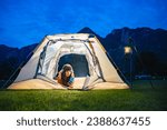 Woman wearing earphones using smartphone to video call with family or friends while lying inside tent. Lamp camping hanging on steel stand. A background of mountain ridge under dark blue sky twilight.