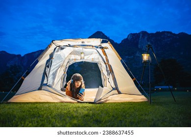 Woman wearing earphones using smartphone to video call with family or friends while lying inside tent. Lamp camping hanging on steel stand. A background of mountain ridge under dark blue sky twilight. - Powered by Shutterstock