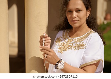 A woman wearing an embroidered white blouse holds prayer beads in a serene outdoor setting. She exudes calm and focus, creating a spiritual and contemplative atmosphere. - Powered by Shutterstock