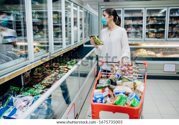 Woman wearing face mask buying in\
supermarket.Panic shopping during Coronavirus covid-19\
pandemic.Budget buying at a supply store.Buying freezer smart\
purchased household pantry\
groceries