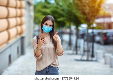 Woman wearing face mask protect from virus showing thumb up. Woman wearing medical face protection mask using smartphone and showing ok sign. - Powered by Shutterstock