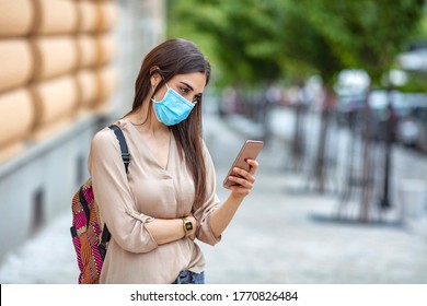 Woman Wearing A Face Mask Walking Outdoors And Using Cell Phone. Woman Using A Phone With The Coronavirus Tracking App. Woman Outdoors Wearing A Facemask While Texting On Her Cell Phone