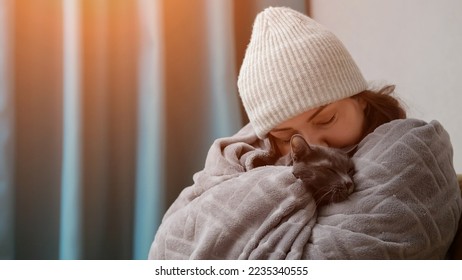 Woman wearing grey hat tries to warm herself and domestic cat with cosy blanket and hugs animal. Adult housewife takes care of domestic pet and shows love, sunlight - Shutterstock ID 2235340555