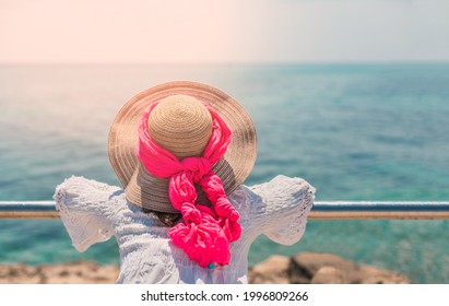 a woman wearing a hat contemplates the blue sea in summer, with her back against a metal railing. the sea is blue and it's summer. welcome summer.