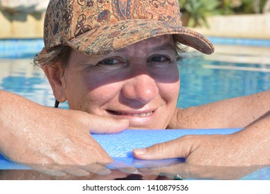 Woman Wearing A Hat, Floating On A Pool Noodle In A Swimming Pool, Smiling To Camera. Summer Vibes, Leisure And Relaxation