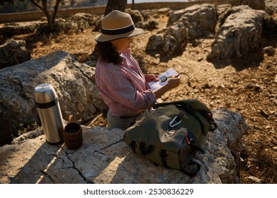 A woman wearing a hat reads a map while sitting on rocks in a sunny, natural setting. A backpack and thermos sit beside her, suggesting a day of adventure and exploration. - Powered by Shutterstock