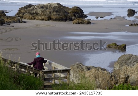 a woman wearing a hat, scarf and jacket starts to walk down a wooden staircase from the top of a large hill to the wet sandy beach below