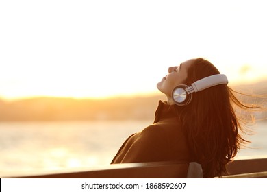 Woman wearing headphones listening to music breathing fresh air relaxing sitting on a bench in winter on the beach - Shutterstock ID 1868592607