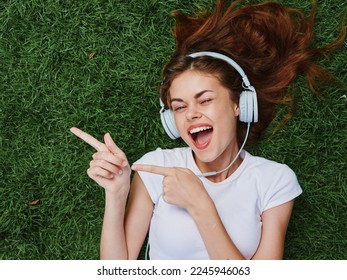 Woman wearing headphones listening to music points the finger to the side, a smile with teeth lies open mouth, lies on the green grass in the summer, good mood happiness
