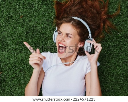 Woman wearing headphones listening to music points the finger to the side, a smile with teeth lies open mouth, lies on the green grass in the summer, good mood happiness