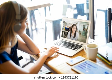 Woman Wearing Headphones And Participating In A Video Conference Call On A Laptop While Telecommuting From A Cafe