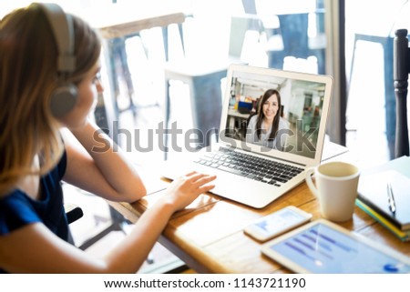 Woman wearing headphones and participating in a video conference call on a laptop while telecommuting from a cafe