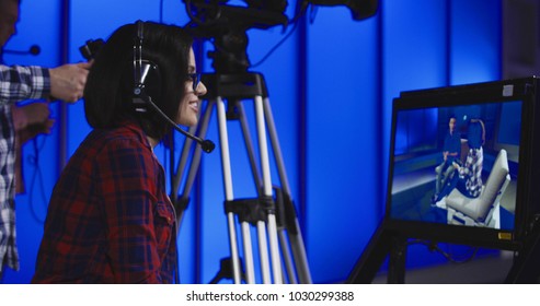 Woman wearing a headset sitting behind a monitor with the cameraman during production giving instructions in a newsroom gesturing and pointing