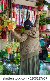 Woman Wearing Hijab And Jacket Hanging The Plant At The Shop Surrounded By Hanging Plants.