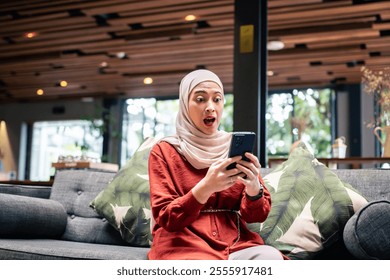 A woman wearing a hijab shows surprise while looking at her smartphone. She sits on a cozy sofa in a modern, well-lit room with a natural background - Powered by Shutterstock