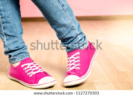 A woman wearing jeans and shocking pink canvas lace-up shoes stands awkwardly, one foot bent inward, pigeon toed.