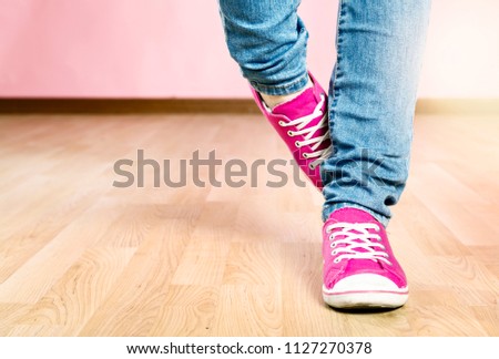 A woman wearing jeans and shocking pink canvas lace-up shoes stands awkwardly, rubbing one foot behind the other.