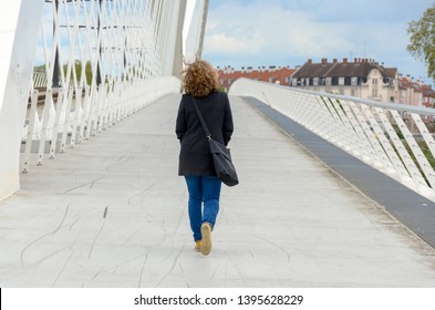 Woman wearing jeans and a warm coat with a large bag over her shoulder walking away across a pedestrian bridge with white receding rails in a town environment