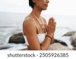 Woman wearing jewelry in prayer pose, enjoying meditation and spiritual moments by the seaside during a beach vacation.