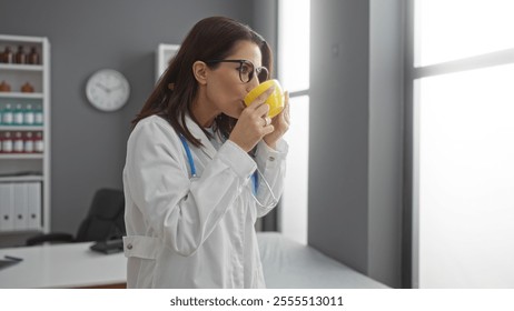 Woman wearing lab coat and glasses sipping from cup in bright clinic room, surrounded by medical supplies and office equipment during workday. - Powered by Shutterstock