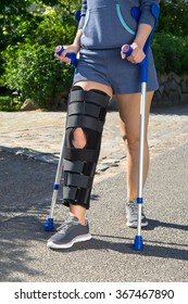 Woman Wearing A Leg Brace With Adjustable Side Panels To Immobilize And Support Her Knee After Surgery Walking On Crutches Outdoors On A Walkway In A Garden