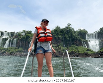 woman wearing life vest standing on boat with Iguazu Waterfalls in background. Powerful cascading waterfalls, lush greenery, and flowing river in natural landscape, Iguazu National Park, Brazil. - Powered by Shutterstock