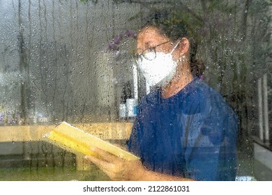 Woman Wearing Mask In Her Quarantine, Reading A Book Behind Wet Window Glass After The Rain