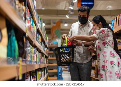 Woman, Wearing A Mask, Places An Item Into A Shopping Basket Being Held By A Man Wearing A Mask.