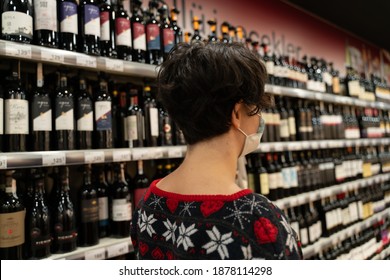 Woman Wearing Medical Mask Choosing Wine At Alcohol Stand. Ugly Christmas Sweater And Shopping For 2021 New Year And Christmas Dinner Table And Home New Year Party. Coronavirus Pandemic. 