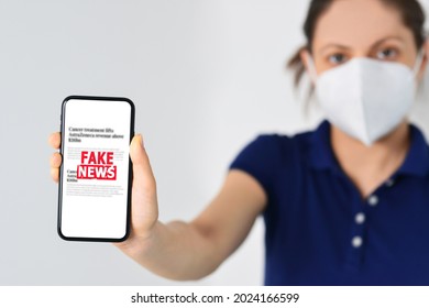 Woman Wearing Medical Mask And Holding A Smartphone With Fake News In Front. Propaganda, Misinformation. Horizontal Photo. Online Fake News On A Mobile Phone. 