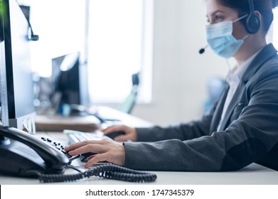 A woman wearing a medical mask in an open space office. A female secretary answers headset clients' calls. Social distance at work. Sanitary standards in the epidemic of coronavirus. - Powered by Shutterstock