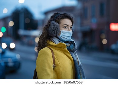 Woman wearing medical protective mask at dusk. Young woman wearing face mask against pollution standing outdoors on winter evening. Girl with earflap in winter city street during coronavirus outbreak.