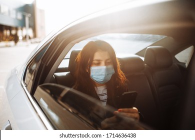 Woman wearing a medical sterile mask in taxi car on a backseat looking out of window checking her cell phone. Girl passenger waiting in a traffic jam during coronavirus quarantine. Healthcare concept