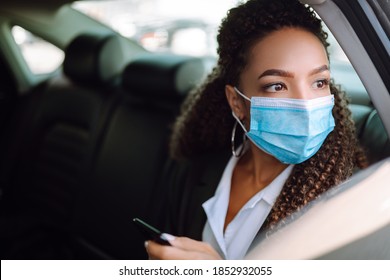 Woman Wearing A Medical Sterile Mask In Taxi Car On A Back Seat Looking Out Of Window Checking Her Cell Phone. Safety And Pandemic Concept. Covid-2019.