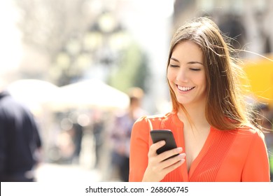 Woman wearing orange shirt texting on the smart phone walking in the street in a sunny day 