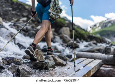 Woman wearing outdoor boots and shorts walking across a wooden bench and rocks crossing a wild alpine creek using hiking poles. Trekking shoes on rocks and in runnig water.