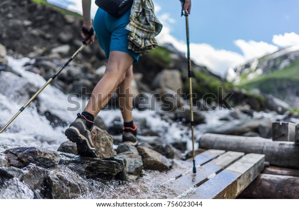Woman wearing outdoor\
boots and shorts walking across a wooden bench and rocks crossing a\
wild alpine creek using hiking poles. Trekking shoes on rocks and\
in runnig water.
