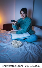 Woman Wearing Pajamas Sitting In Bed, Eating Popcorn And Laughing While Watching Funny Movie On TV, Relaxing At Home Late At Night