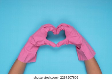 woman wearing pink cleaning gloves on light blue background making heart sign with hand - Shutterstock ID 2289507803