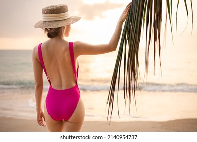 Woman wearing pink one piece swimsuit and straw hat enjoy romantic sunset moment under the coconut trees on tropical beach.